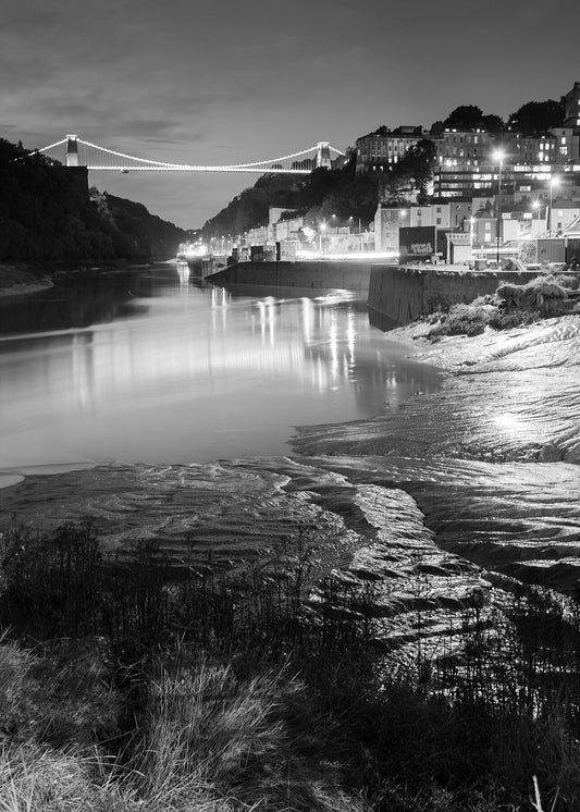 This black and white photograph shows the view of the Avon Gorge towards Clifton and the suspension bridge at night, with lights reflected on the water