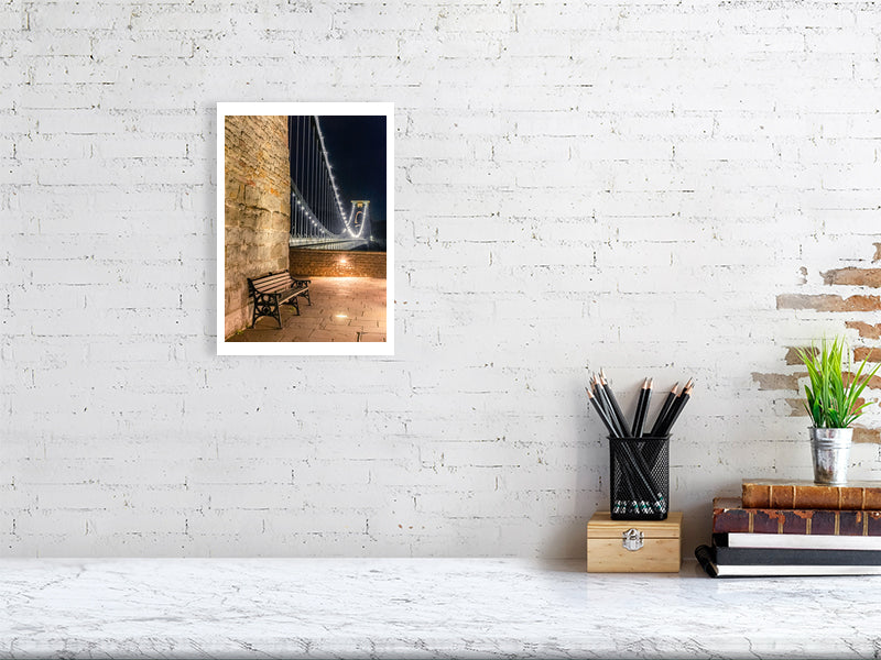 À brick wall with a central print of Clifton suspension bridge in Bristol at night with with a foreground seat and sparkling lights