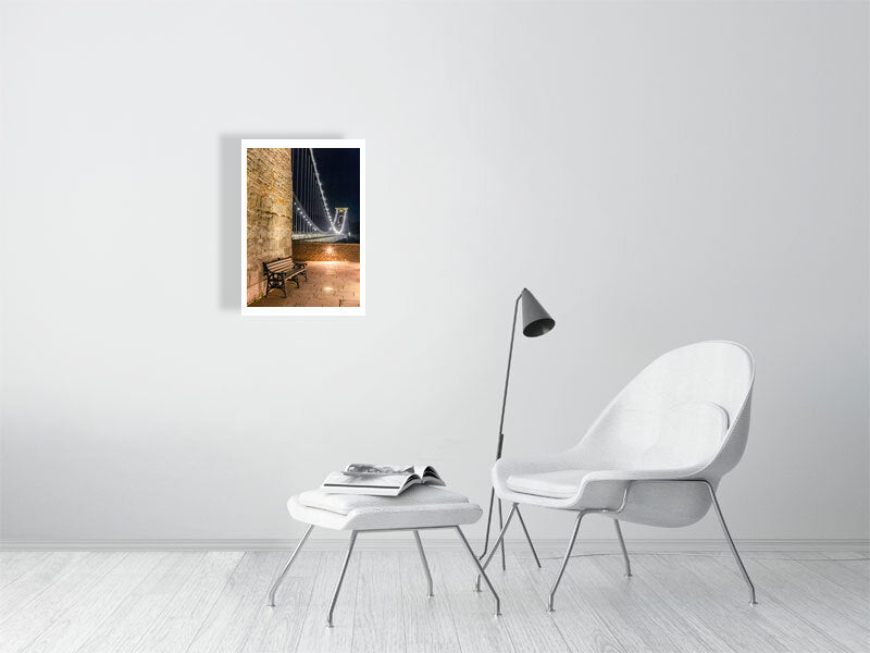 A white brick wall with a hanging picture of Clifton suspension bridge in Bristol at night with with a foreground seat and sparkling lights