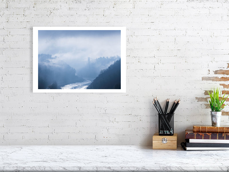 A white brick wall with a hanging picture of Clifton Suspension Bridge in the distance breaking free of the morning fog, with the River Avon below and the Avon gorge