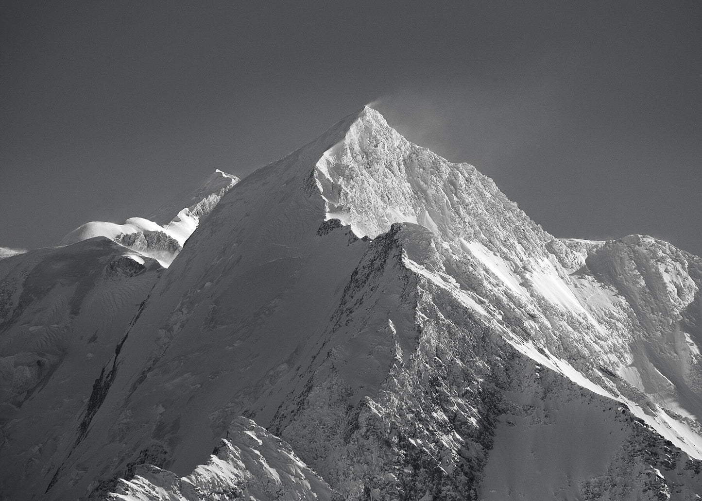 Black and white high res photo of the Aiguille du Bionnassay forms a dramatic ridge-line to the summit of Mt Blanc high above Chamonix, seen just behind in the distance as the strong summit winds lift snow from the pea