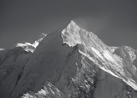 Black and white high res photo of the Aiguille du Bionnassay forms a dramatic ridge-line to the summit of Mt Blanc high above Chamonix, seen just behind in the distance as the strong summit winds lift snow from the pea