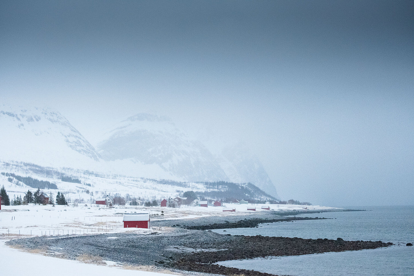 Red Norwegian hut on the shoreline of the Fjord while snow swaps in across Kvaloya island