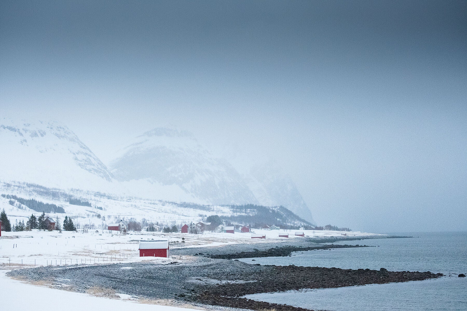 Red Norwegian hut on the shoreline of the Fjord while snow swaps in across Kvaloya island