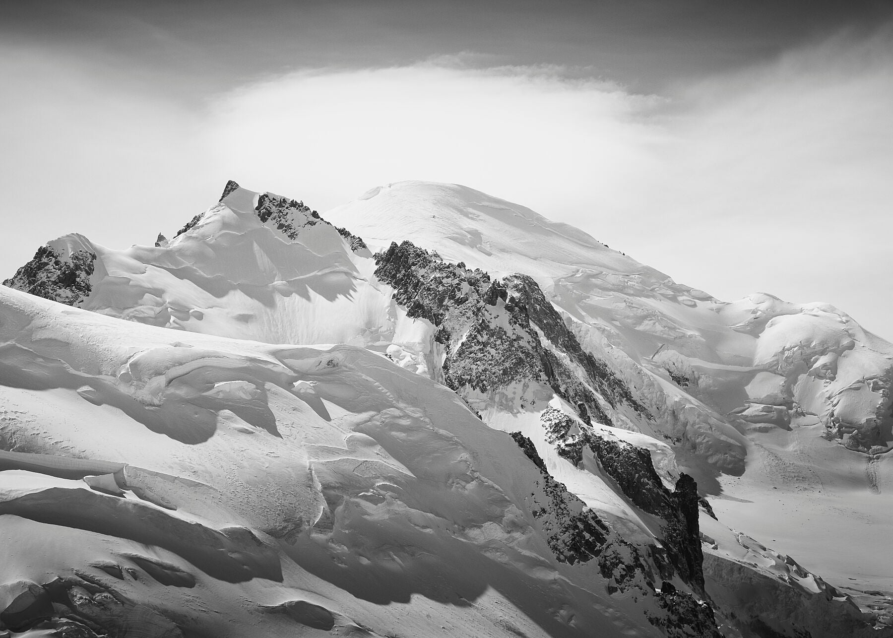 A stunning high resolution black and white photograph of summit of Mont Blanc capped by a lenticular cloud, emerging from behind the summit of the foreground Mt Tacul and glacial landscape of the Mont Blanc Massif.