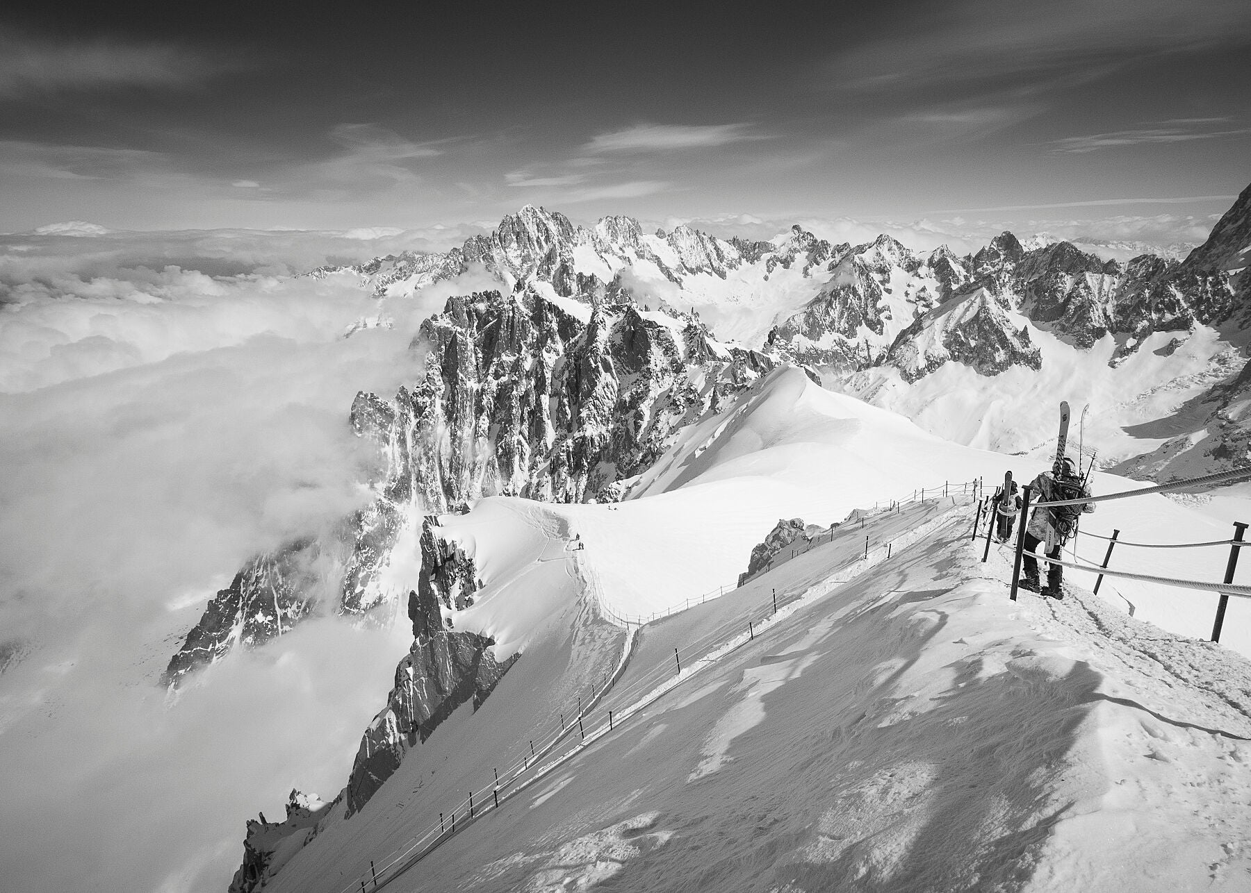 A black and white high resolution photograph  of skiiers walking down the arête at the start of the Vallee blanche off piste route from the Aiguille du Midi station in the Mont Blanc Massif. The image captures the precipitous nature of the aerate, a formidable obstacle for nervous first timers to navigate before the ski home can commence.