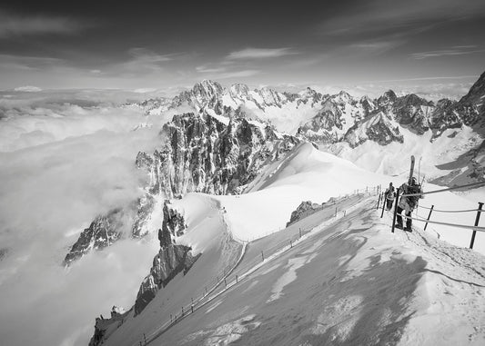 A black and white high resolution photograph  of skiiers walking down the arête at the start of the Vallee blanche off piste route from the Aiguille du Midi station in the Mont Blanc Massif. The image captures the precipitous nature of the aerate, a formidable obstacle for nervous first timers to navigate before the ski home can commence.