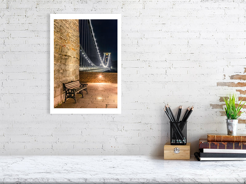 À white brick wall with a hanging photograph of Clifton suspension bridge in Bristol at night with with a foreground seat and sparkling lights