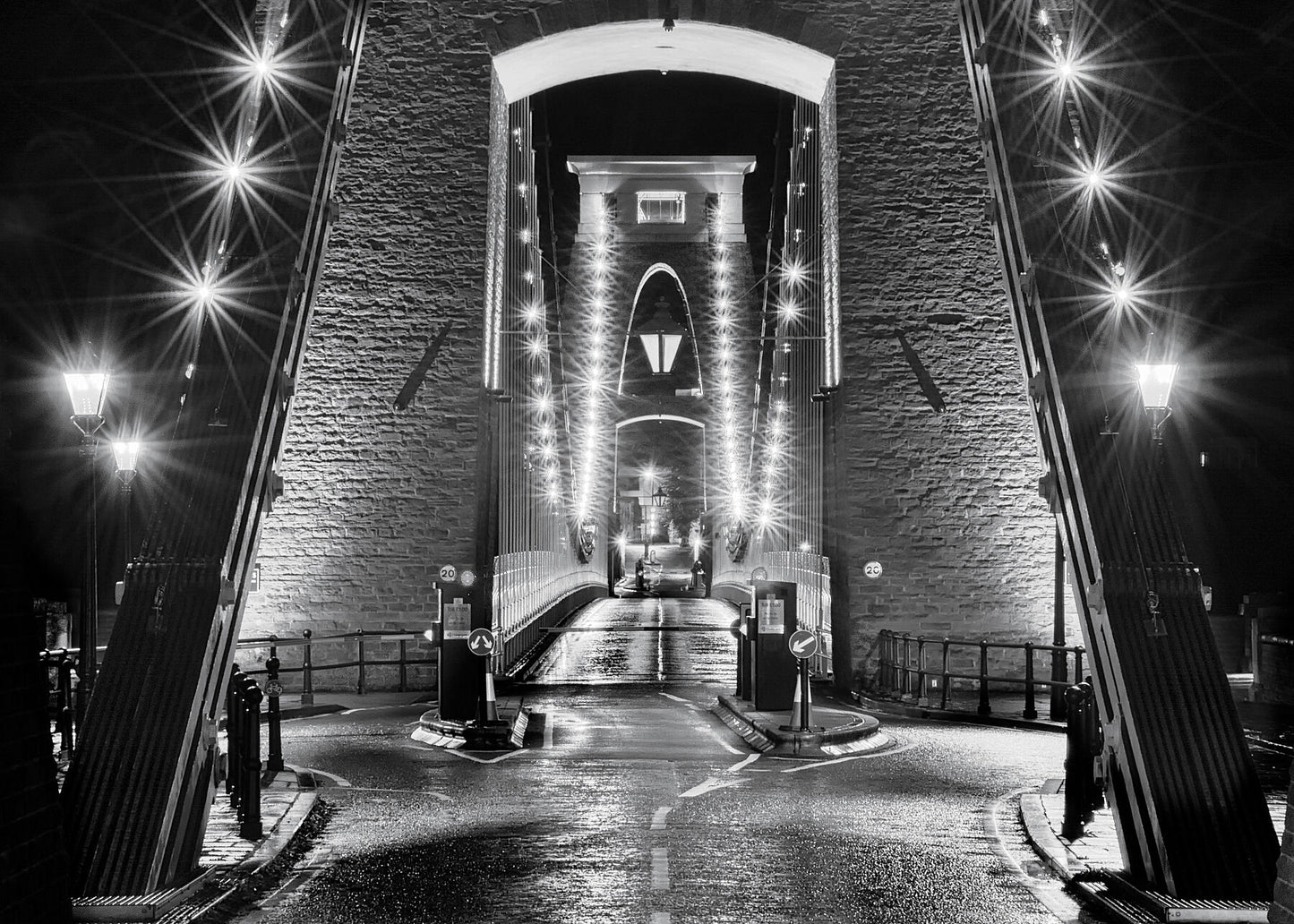Unusual and dramatic black and white fine art photograph of Brunel’s Clifton suspension bridge in central Bristol at night  with light reflections on the wet tarmac