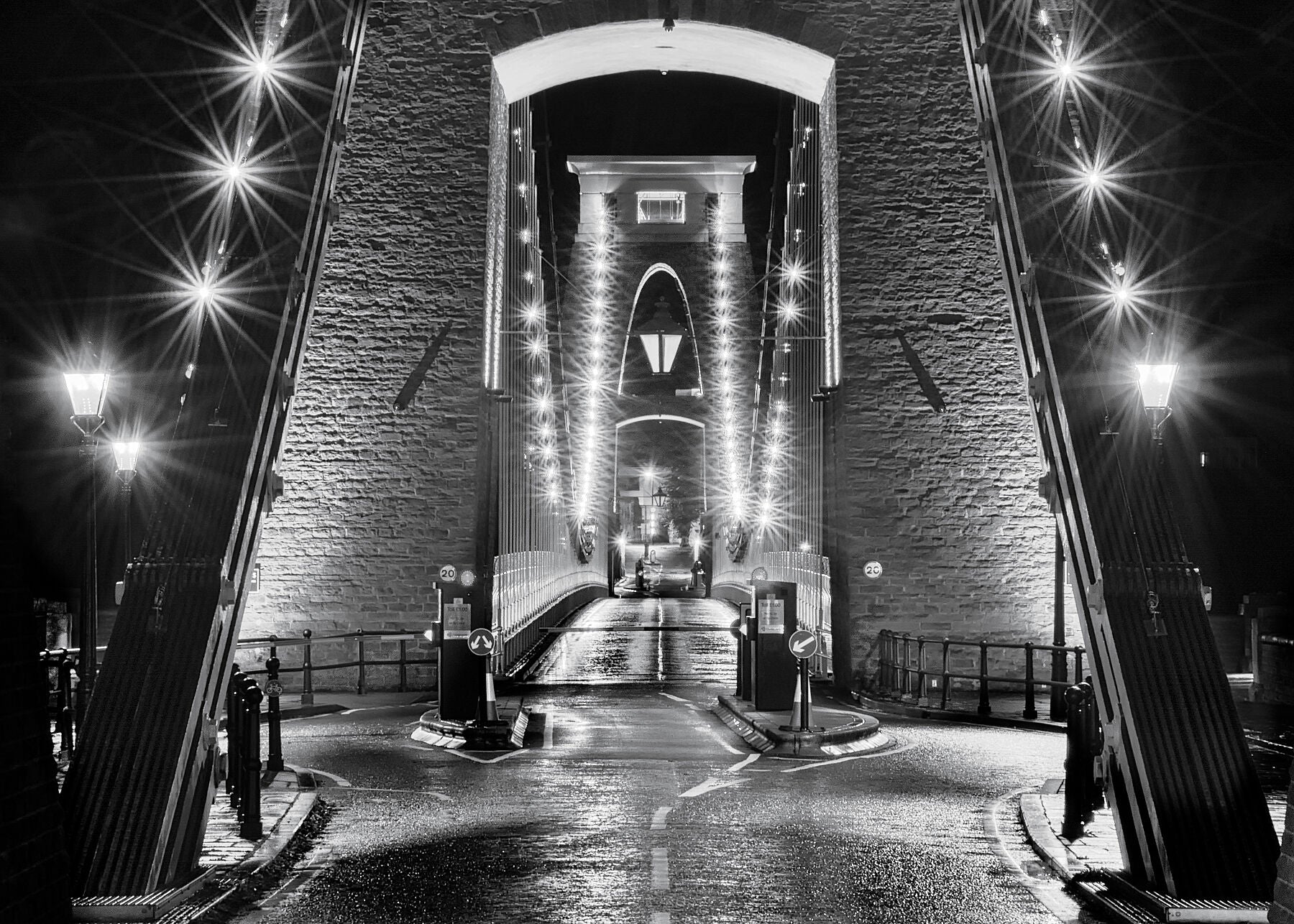 Unusual and dramatic black and white fine art photograph of Brunel’s Clifton suspension bridge in central Bristol at night  with light reflections on the wet tarmac