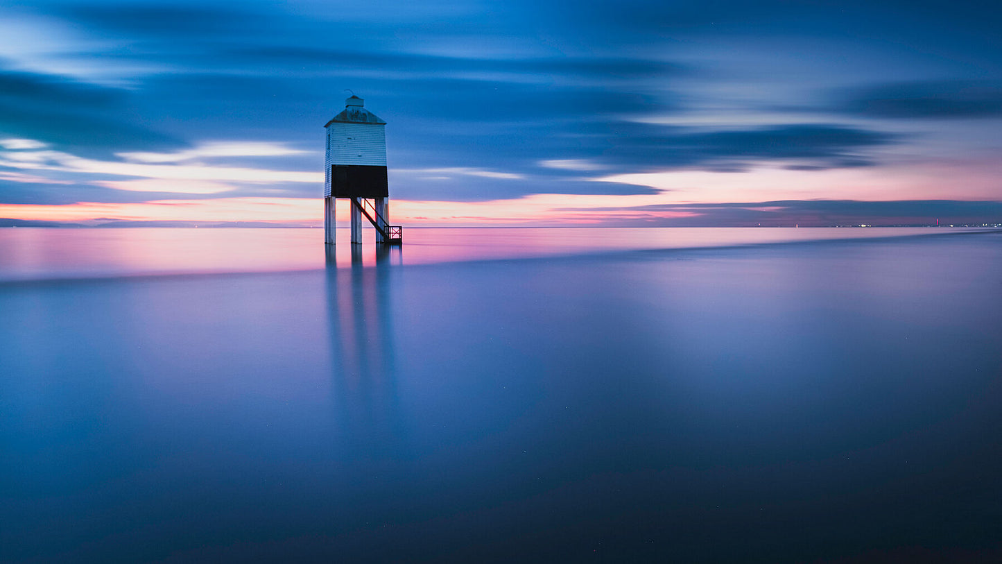 The sun sets behind the Burnham Low Lighthouse in the Severn estuary, reflecting golden hues in the tranquil waters as the tide sweeps inshore. A tranquil and calming photograph
