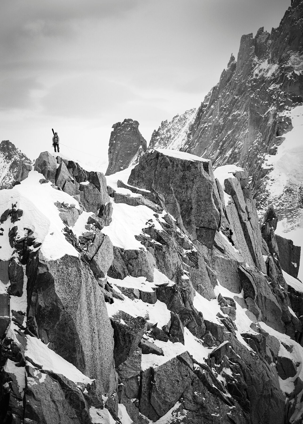 À climber with skis on his back stands at the top of a jagged ridge line in tje Mont Blanc massif