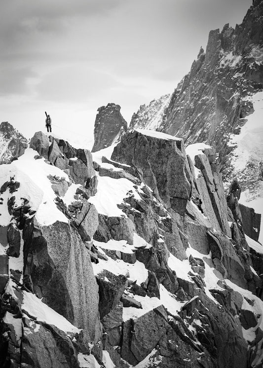 À climber with skis on his back stands at the top of a jagged ridge line in tje Mont Blanc massif