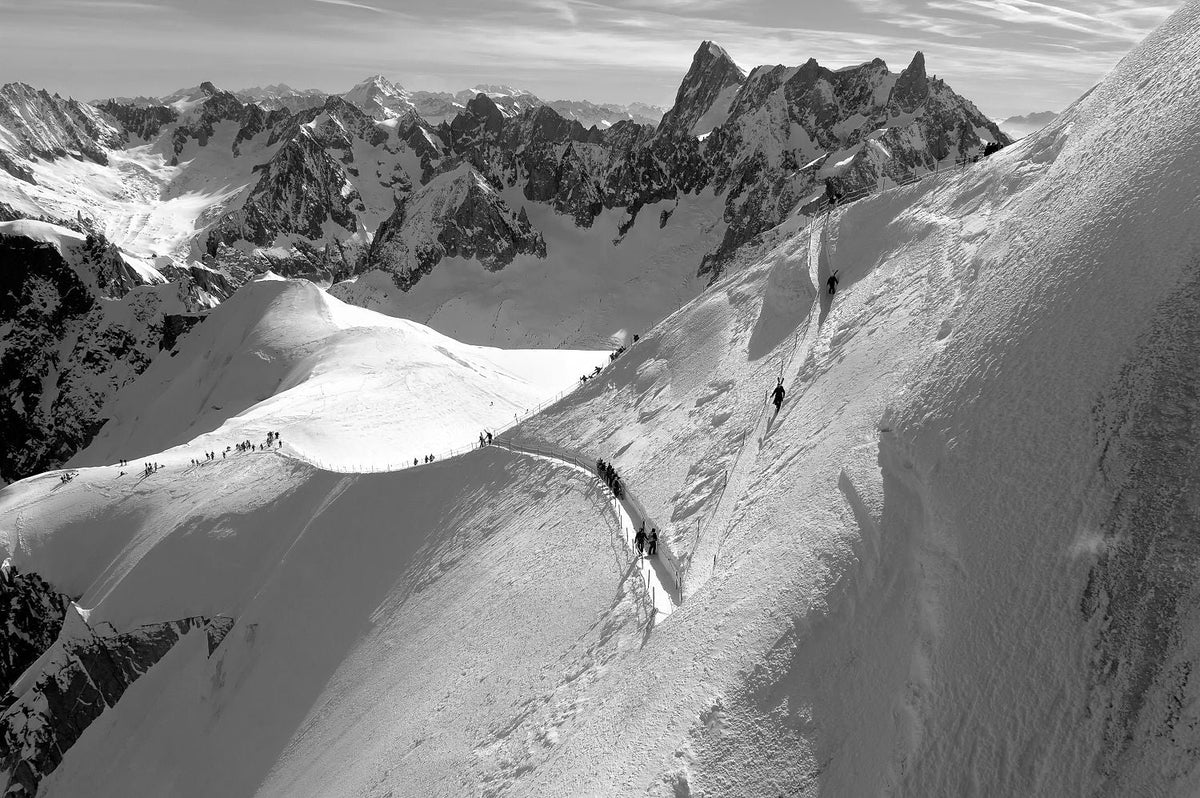Black-and-white photograph of skiers descending the snowy Aiguille du Midi arête above Chamonix, French Alps.