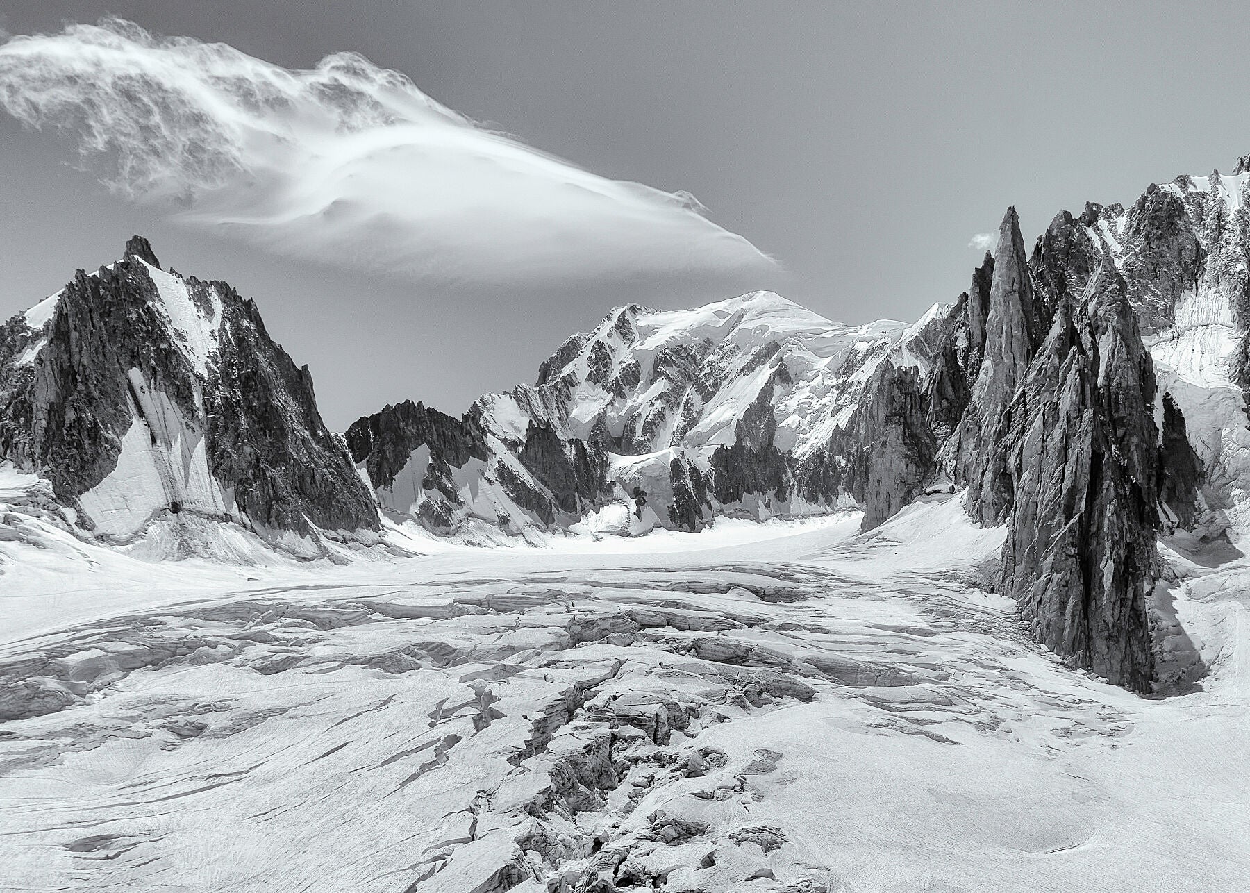 Black and white photograph capturing the view over a glacier towards the summit of Mont Blanc, with dramatic peaks and a beautiful cloud