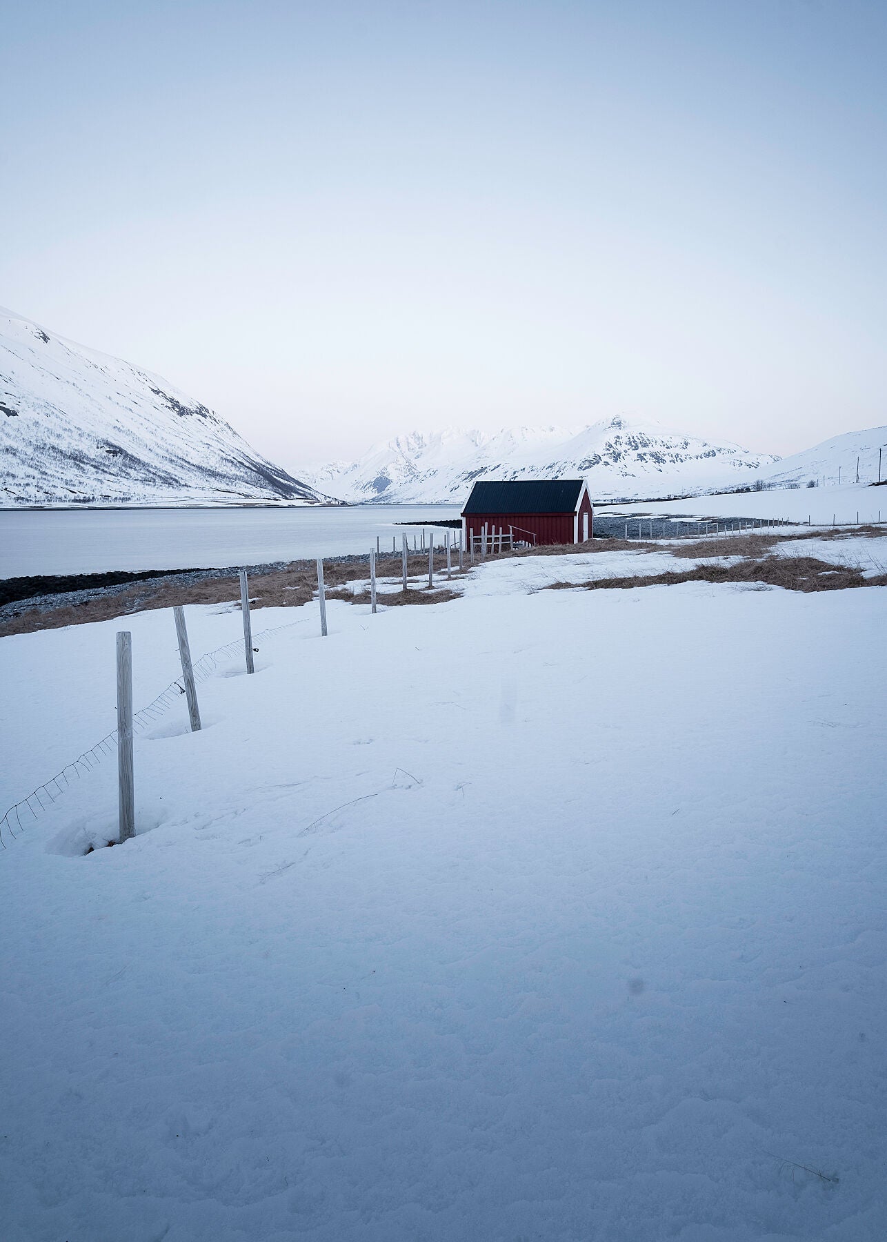 High res photograph of a fence leading towards a fishermans hut in snowy Norway, with Mountain and a fjord in the distance